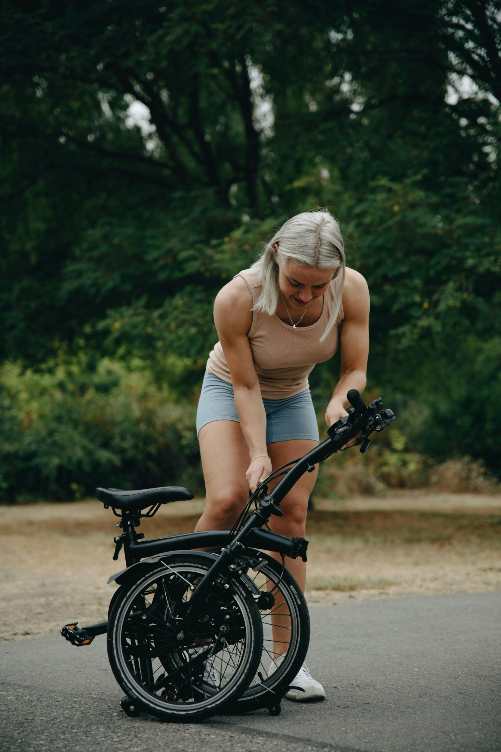 kobieta, blondynka o umięśnionych ramionach składa rower elektryczny nachylając się nad nim, manewruje dłonią przy kierownicy, rower jest w kolorze czarnym.